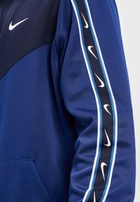 Blå Nike-tröja med en mörkblå övre del, vitt logotyp och sidostråk med ljusblå och svarta accenter. Mjuk tygtextur.