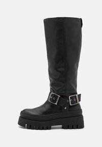 Botas altas de cuero negro con suela de plataforma gruesa, que presentan hebillas plateadas y una superficie texturizada. Costuras laterales visibles, acabado mate.