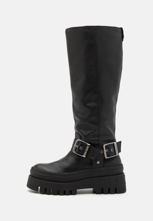 Bottes noires en cuir long avec une semelle épaisse, dotées de boucles en argent et d'une surface texturée. Coutures latérales visibles, finition mate.