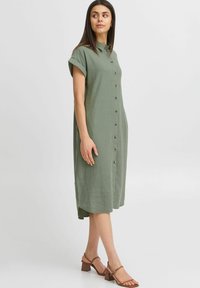 Abito camicia a maniche corte di colore verde oliva, realizzato in tessuto leggero. Caratterizzato da maniche a cappuccio, colletto e una silhouette che arriva fino al ginocchio con bottoni frontali.