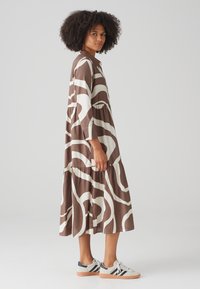 Brown long dress with white abstract patterns, featuring a tiered design and three-quarter sleeves. Paired with multi-colored athletic sneakers.