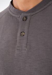 Graues Baumwoll-Henley-Shirt mit Rundkragen, zwei Knöpfen und subtiler Struktur. Die Knöpfe sind hellfarben und haben eine leicht glänzende Oberfläche.