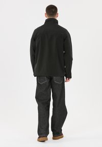 Svart jacka med hög krage, kombinerad med mörka jeans i denim som har synliga bakfickor och en ljusare söm. Bruna skor med gummisula.