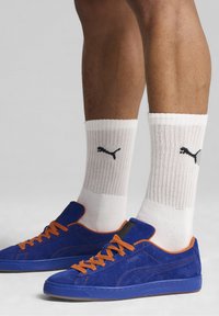 Niebieskie zamszowe sneakersy z pomarańczowymi sznurowadłami, charakteryzujące się fakturowaną powierzchnią i czarnymi akcentami logo, noszone z białymi skarpetami w prążki.