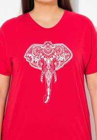 T-shirt rouge avec un graphique d'éléphant blanc présentant des motifs complexes et des détails floraux, col rond et manches courtes.