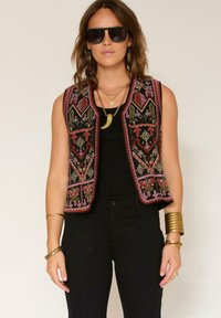 Gilet noir brodé avec des motifs floraux vibrants en rose, vert et rouge, associé à un haut noir et des accessoires dorés.