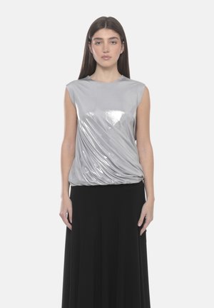 Donna con top argento lucido senza maniche e gonna lunga nera, in piedi con le braccia rilassate contro uno sfondo bianco.