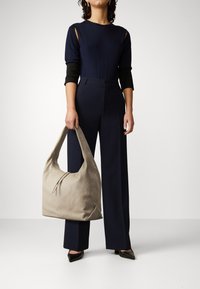 Beige läder hobo-väska med en mjuk, slapp form, med dragkedjestängning och bred axelrem, hålls av en person i en mörk outfit.