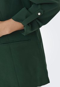 Grön kavaj med uppvikt ärm som har en pärlaccent, slät textur och en detaljerad sidoficka. Skräddarsydd form för en åtsittande look.