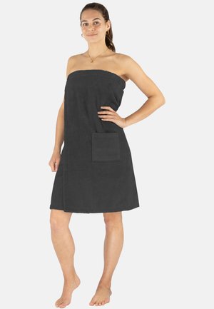 Dunkelgraues Handtuchkleid aus weichem, strukturiertem Stoff. Verfügt über ein trägerloses Design und eine Fronttasche. Wird barfuß in entspannter Pose getragen.