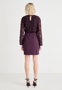 Lila klänning med en texturerad, broderad spetsöverdel med långärmar, nyckelhål i ryggen och en figurnära kjol. Svarta klackar med remmar.