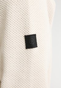 Tessuto di colore crema con una texture a nido d'ape, con una etichetta a patch quadrata nera con logo e testo in rilievo.