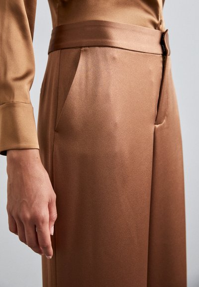 Vue à hauteur de taille de pantalons bruns doux, semblables à du satin, avec une texture lisse, présentant une poche latérale et un devant plat, associés à un chemisier assorti.