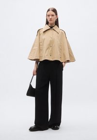 Bež kratka kabanica s velikim ovratnikom, kopčama na pritisak i bočnim džepovima, uparena s crnim širokim hlačama i malom torbicom.