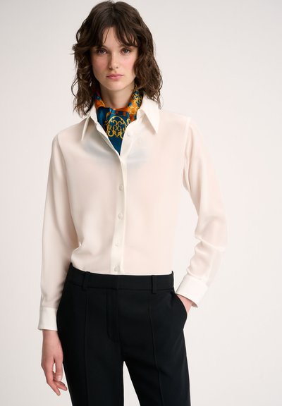 White long-sleeve blouse with a button-down front and a classic collar, paired with black tailored pants. Scarf features an intricate blue and gold pattern.