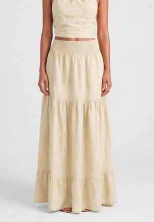 Vrouw draagt een beige, hooggetailleerde, gelaagde maxi-rok gecombineerd met een bijpassend cropped mouwloos topje, staand tegen een effen achtergrond.