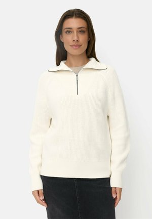 Ung kvinde iført en hvid strikket sweater med halv lynlås i kraven og et sort skørt, stående mod en ensfarvet lys baggrund.
