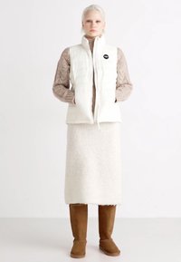 Chaleco acolchado blanco sobre un suéter de punto marrón claro, combinado con una falda blanca de felpa y botas marrones hasta la rodilla. Diseño simple y texturas suaves.