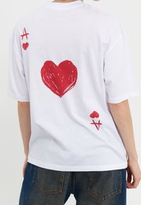 Vit t-shirt med kort ärm som har en stor röd hjärtgrafik på baksidan, tillsammans med en röd diamant- och spader-symbol ovanför.