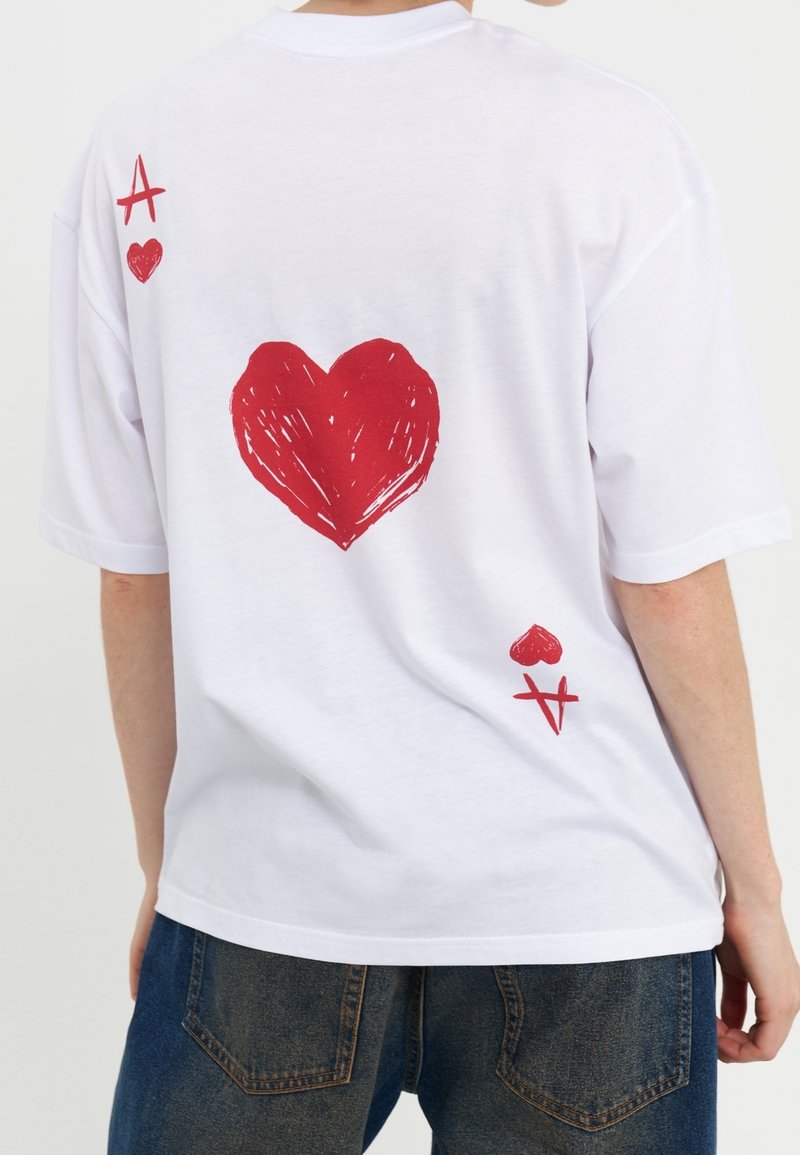 Vit t-shirt med kort ärm som har en stor röd hjärtgrafik på baksidan, tillsammans med en röd diamant- och spader-symbol ovanför.