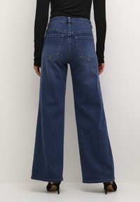 Jeans in denim blu a gamba larga con vita alta, dotati di tasche posteriori e orlo pulito. Abbinati a scarpe nere con tacco alto.