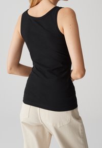 Personne mince portant un haut noir sans manches et un pantalon beige clair, de dos, les mains derrière le dos, sur un fond neutre.