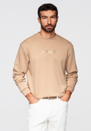Man met baard, gekleed in een beige sweatshirt met de tekst "EXIMIO" en witte broek, handen in de zakken, staand tegen een effen lichte achtergrond.