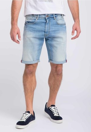 Mann trägt hellblaue hochgekrempelte Jeansshorts und schwarze Sneakers mit weißen Schnürsenkeln, steht vor einem schlichten Hintergrund.