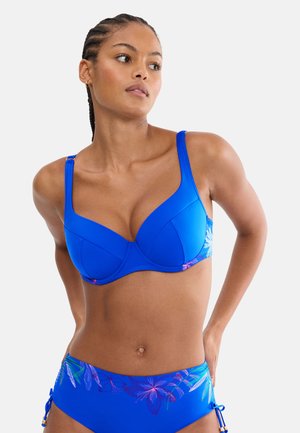 Vrouw met gevlochten haar die een blauwe bikini draagt met bloemmotieven aan de zijkanten en verstelbare bandjes, staand tegen een effen achtergrond.