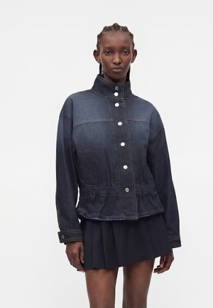Kvinde med fletninger iført en mørk denimjakke med hvide knapper og et sort plisseret skørt mod en ensfarvet hvid baggrund.