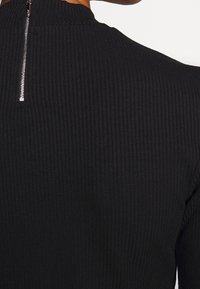 Czarny, prążkowany top z wysokim kołnierzem i zapięciem na zamek z tyłu. Materiał ma teksturowany, elastyczny wygląd.