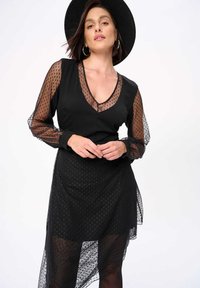 Zwarte jurk met lange doorschijnende stippenmouwen en een gelaagde asymmetrische zoom. Beschikt over een V-halslijn en een gestructureerde stof.