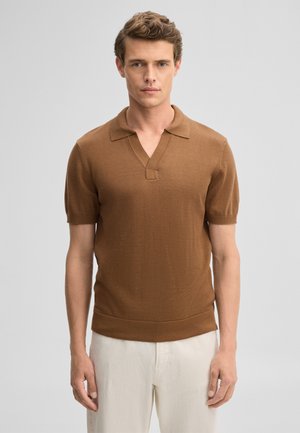 Junger Mann mit kurzen lockigen Haaren, der ein braunes kurzärmeliges Strick-Poloshirt und helle beige Hosen trägt, steht vor einem schlichten Hintergrund.