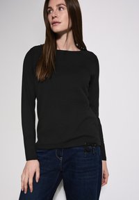 Musta pitkähihainen paita, jossa on teksturoitu kudos, pyöreä pääntie ja säädettävä nyöri helmassa, yhdistettynä tummansinisiin farkkuihin.