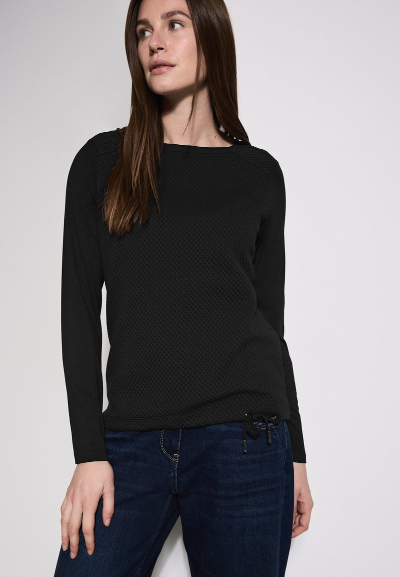 Musta pitkähihainen paita, jossa on teksturoitu kudos, pyöreä pääntie ja säädettävä nyöri helmassa, yhdistettyn�ä tummansinisiin farkkuihin.