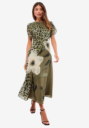 Vrouw die zelfverzekerd loopt in een groene midi-jurk met luipaardprint en grote witte bloemenpatronen, met zwarte schoenen met enkelbandje.
