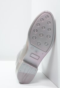 Semelle de chaussure transparente avec une surface texturée, présentant des clous de traction circulaires et une plaque logo rectangulaire, le tout dans des couleurs atténuées.