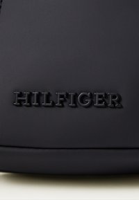 Fekete matt táska, a "HILFIGER" logó dombornyomott felirattal, letisztult, elegáns dizájnnal, sima felülettel és minimális hardver részletekkel.