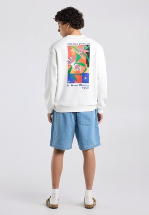 Persoon met krullend haar draagt een wit sweatshirt met kleurrijke Henri Matisse "Creole Dancer" print, denim shorts, witte sokken en sandalen.