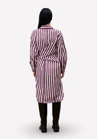 Frau mit langen schwarzen Haaren, die ein rosa und bordeauxrotes, vertikal gestreiftes Kleid und schwarze kniehohe Stiefel trägt, steht mit dem Rücken zugewandt.