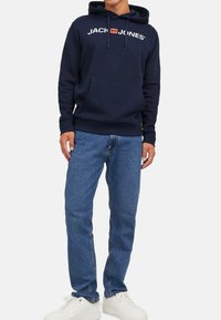 Marineblauer Hoodie mit einer vorderen Kängurutasche und Kordeln, ausgestattet mit einem weißen Logo. Kombiniert mit hellblauen Jeans und weißen Sneakern.
