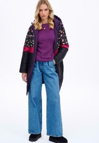 Dlouhý černý péřový kabát s květinovými a vínovými panelovými akcenty, nošený přes fialový svetr a světlé modré volné džíny, doplněný černými botami.