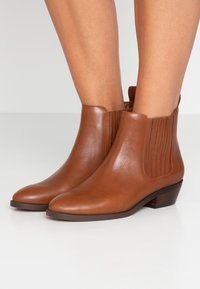 Botines marrones de cuero con paneles laterales elásticos, punta afilada y tacón bajo tipo bloque. Textura lisa con detalles mínimos de costura.