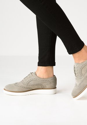 Persoon die lichtgrijze suède brogue schoenen draagt met witte zolen en een zwarte slim fit broek, zijaanzicht met één voet licht opgeheven.