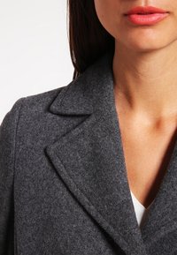 Cappotto grigio in misto lana con rever a punta, tessuto testurizzato e vestibilità sartoriale, con una maglietta a V visibile sotto.