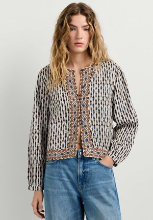Junge Frau mit welligem blonden Haar, die eine gemusterte, geknöpfte Bluse mit langen Ärmeln und blaue Jeans trägt, eine Hand in der Tasche.