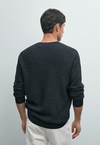 Mann mit dunklen Haaren, der einen dunkelgrauen Pullover und weiße Hosen trägt, vor einer hellgrauen Wand steht, mit entspannten Armen an den Seiten.