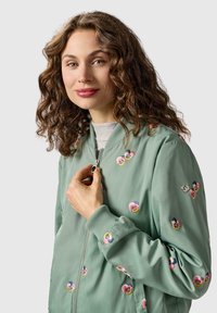 Młoda kobieta z kręconymi włosami, ubrana w jasną zieloną bomberkę z kwiatowym haftem, trzymająca suwak prawą ręką.