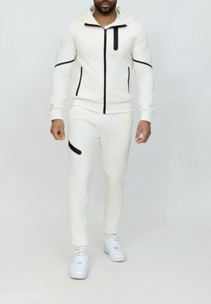 Ensemble en polaire blanc avec fermeture éclair noire et accents, comprenant une capuche, des manches longues et un pantalon ajusté avec une poche latérale. Baskets blanches.