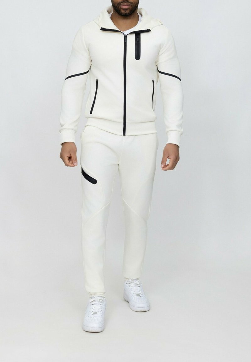 Ensemble en polaire blanc avec fermeture éclair noire et accents, comprenant une capuche, des manches longues et un pantalon ajusté avec une poche latérale. Baskets blanches.